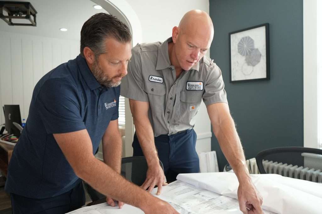 The Werrlein Companies employees plan a plumbing or HVAC solution for a client. Both men are hunched over a table and talk while working on a master plan.